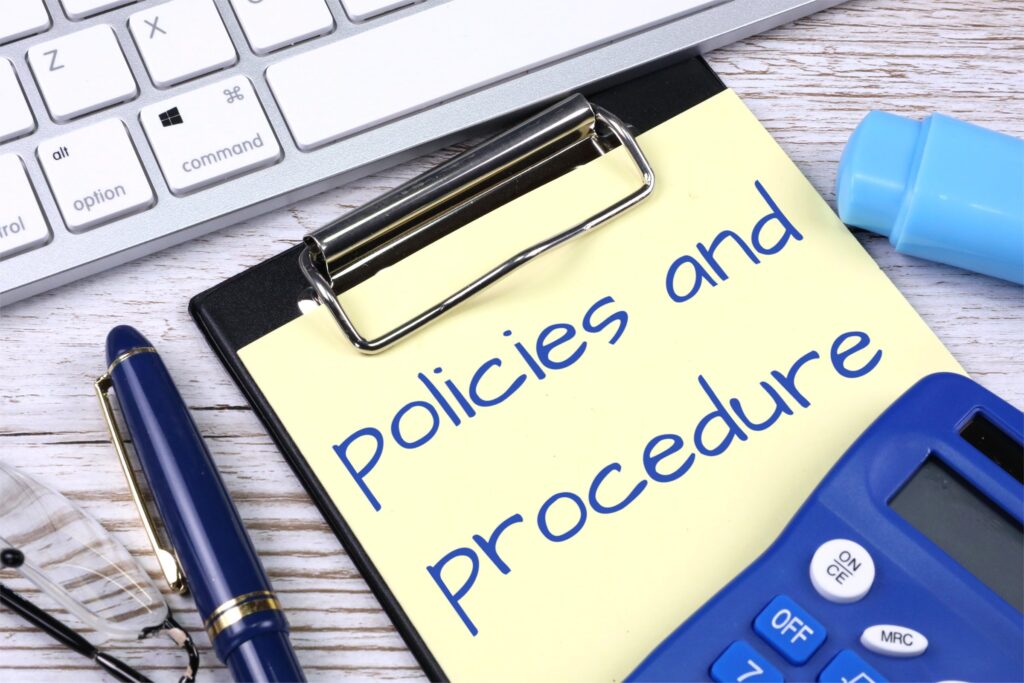 Part of a keyboard, a pen and a clipboard with a post it stating Policies and Procedure